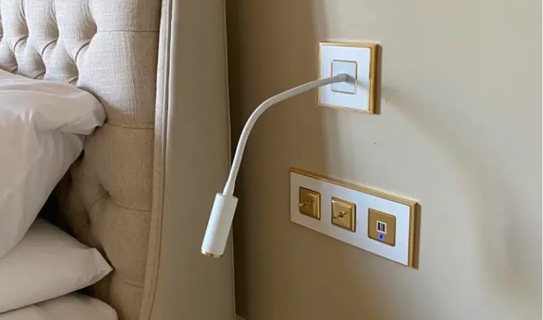 Bedside lighting setup with a flexible white plug-in adaptor cable emerging from a wall socket and gold-accented switches on a beige wall. Bedside lighting setup with a flexible white plug-in adaptor cable emerging from a wall socket and gold-accented switches on a beige wall.