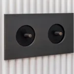 Black dual round-wall switch plate with two exposed brass toggles on a brushed surface mounted on a ribbed wall.