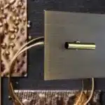 Brass wall switch with a brushed square plate and small cylindrical toggle against a textured dark backdrop.