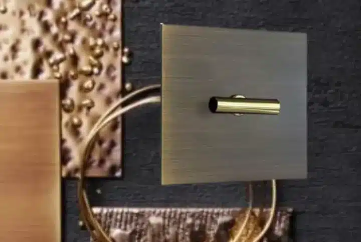 Brass wall switch with a brushed square plate and small cylindrical toggle against a textured dark backdrop.
