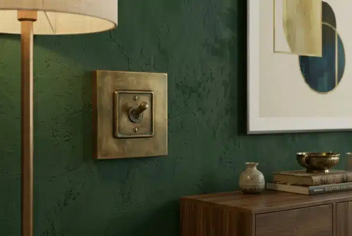 Brass wall switch with square plate mounted on a textured dark green wall, beside a wooden cabinet and abstract framed art.