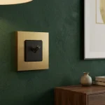 Brushed brass light switches uk: a square brass wall plate with a black toggle switch mounted on a dark green wall, next to a wooden cabinet.
