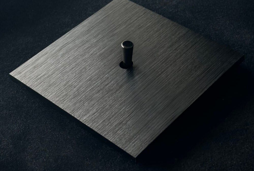 Brushed metal square plate with a small cylindrical black knob in the center, on a dark surface.