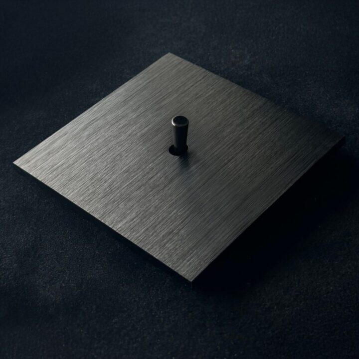 Brushed metal square plate with a small cylindrical black knob in the center, on a dark surface.