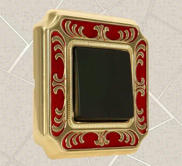 Luxurious brass lighting switch with red enamel detailing and ornate gold scrollwork, mounted on a tiled wall. Luxurious brass lighting switch with red enamel detailing and ornate gold scrollwork, mounted on a tiled wall.