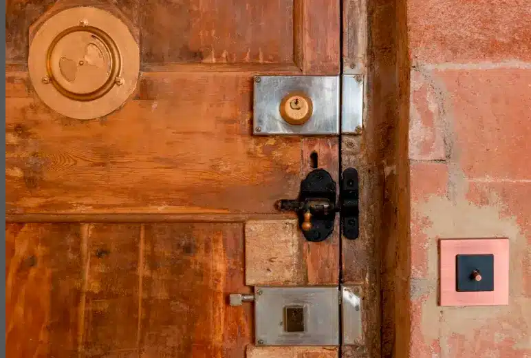 Old wooden door with brass lock and metal plates; a pink-framed light switch on the right wall. Old wooden door with brass lock and metal plates; a pink-framed light switch on the right wall.