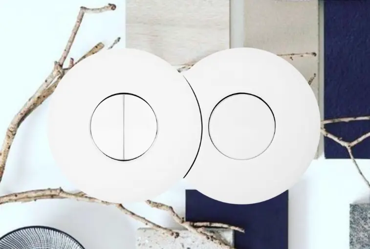 Two white circular wall aluminium switches with central black-ring detailing, set against a collage backdrop of natural branches and coloured panels. Two white circular wall aluminium switches with central black-ring detailing, set against a collage backdrop of natural branches and coloured panels.