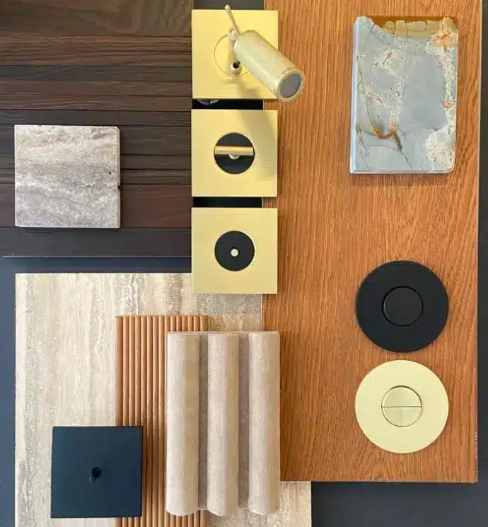 Brass and black modern light switch panels and decorative fixtures arranged on wooden and stone boards, including cylindrical knob, square plates, and fabric samples. Brass and black modern light switch panels and decorative fixtures arranged on wooden and stone boards, including cylindrical knob, square plates, and fabric samples.