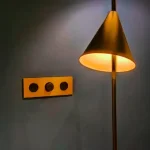 A brass wall plate with three circular sockets beside a warm-lit brass floor lamp against a grey wall.