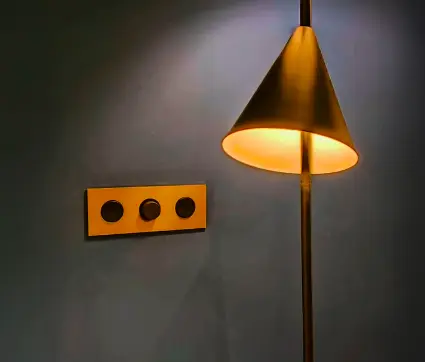 A brass wall plate with three circular sockets beside a warm-lit brass floor lamp against a grey wall.