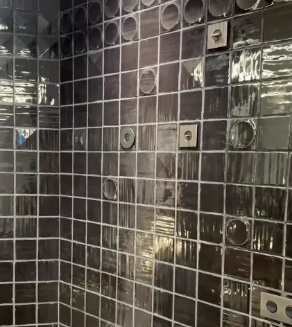Dark tiled wall with small recessed squares and circular fixtures; metallic, reflective surface. Dark tiled wall with small recessed squares and circular fixtures; metallic, reflective surface.