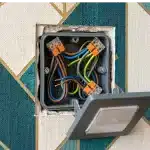 Electrical light switch box with exposed wiring and a tilted, open faceplate mounted in a geometric-patterned wall.