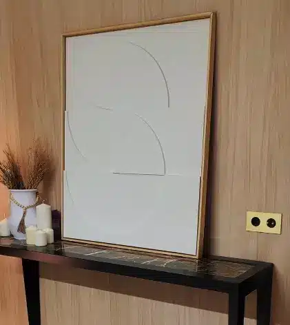 Framed white abstract wall art with grey curved lines leaning on a black console table by a wooden wall. Candles and a vase sit nearby.