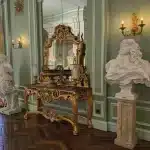 Luxurious antique-style entry with ornate gold console table, large gilded mirror, marble busts, and classical wall sconces in a green-paneled room. Luxury light fixture brands context.