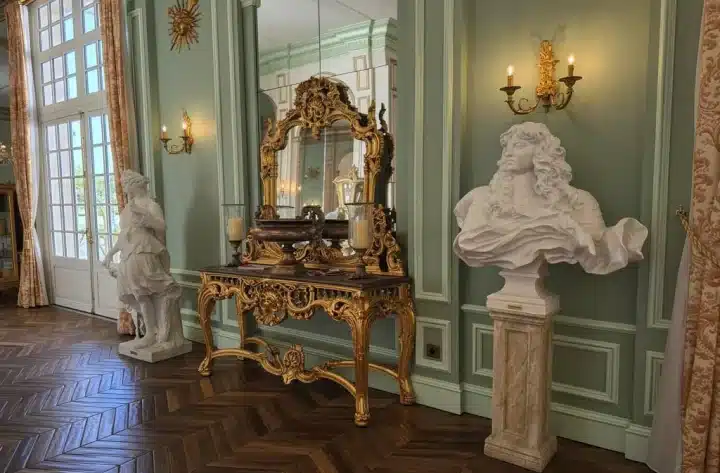 Luxurious antique-style entry with ornate gold console table, large gilded mirror, marble busts, and classical wall sconces in a green-paneled room. Luxury light fixture brands context.