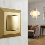 Luxury interior switches design: gold-toned wall light switch near a marble wall, with a stylish living room in the background.
