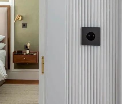 Modern bedroom doorway with a built-in grey wall panel and a black circular electrical outlet; bedside table visible in the adjoining room. Modern bedroom doorway with a built-in grey wall panel and a black circular electrical outlet; bedside table visible in the adjoining room.