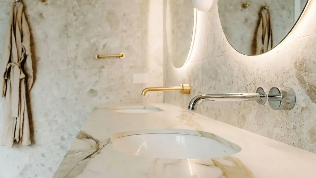 Shiny chrome and brass bathroom taps mounted on a marble countertop, with a circular mirror and soft lighting in a modern, elegant washroom. difference between chrome and brass taps integrated.
