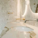 Shiny chrome and brass bathroom taps mounted on a marble countertop, with a circular mirror and soft lighting in a modern, elegant washroom. difference between chrome and brass taps integrated.