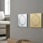 Two wall-mounted recessed socket plates: one silver chrome and one brass, both with circular covers, on a grey wall above a wooden surface.